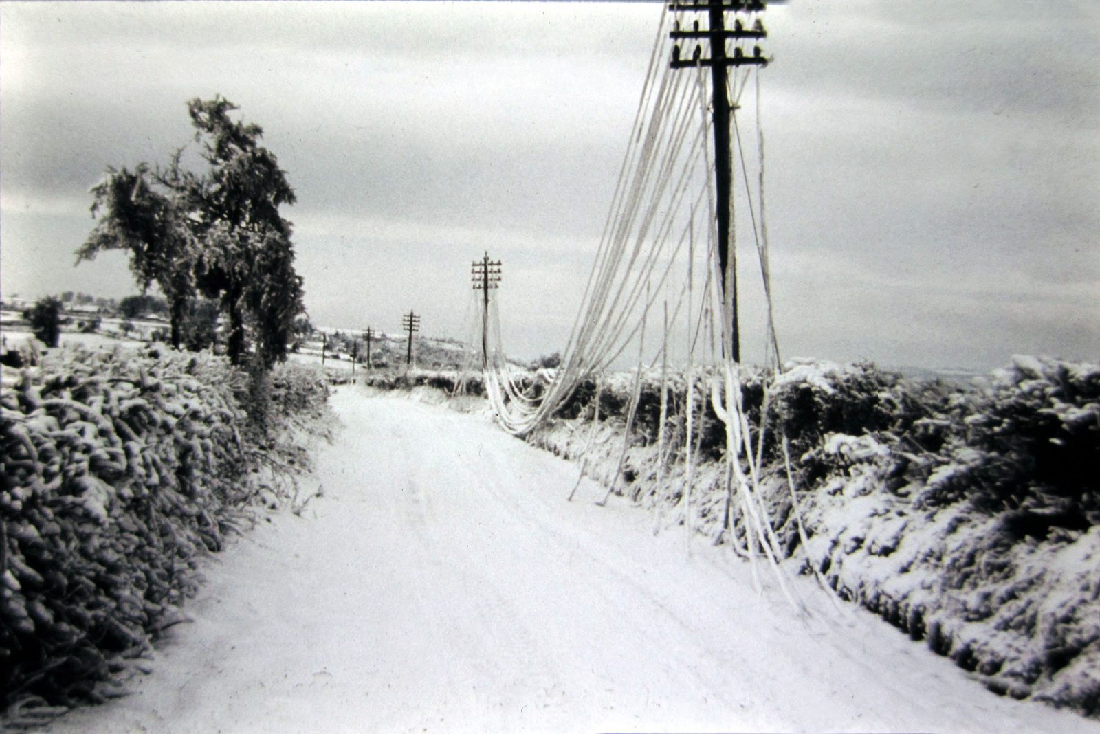 Vintage black and white photograph of a snow-covered UK country lane with overhead telephone poles and wires heavily bowed under the weight of ice and snow — UK Prepper Hub