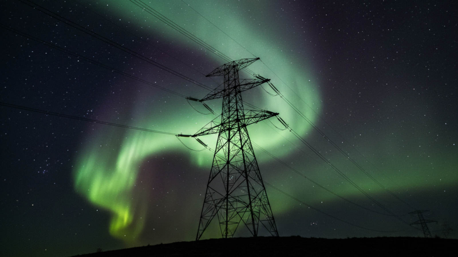 Silhouette of a UK power pylon under a vibrant green and purple Aurora Borealis sky, representing solar storm risks to the grid.