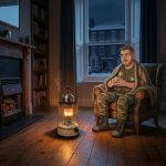 Cartoon Keith with a mug of tea beside a lit paraffin heater with a kettle on top in a dark UK living room during a power cut